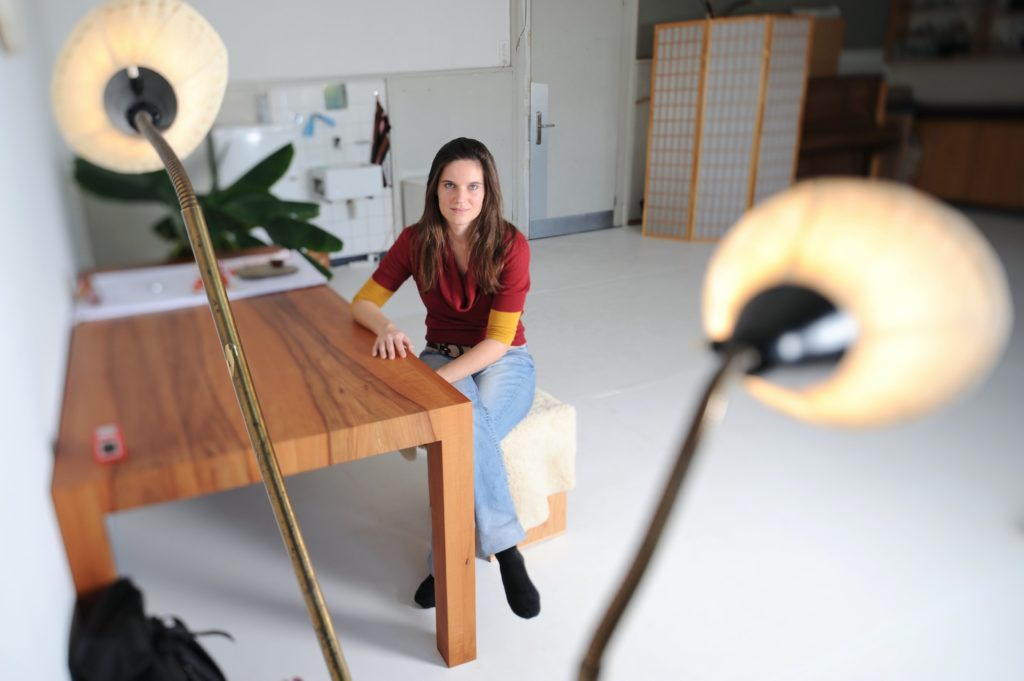 Eine Frau sitzt an einem Holztisch, die rechte Hand darauf gelegt, die Beine verschränkt und den linken Arm darauf gelegt. Im Vordergrund rechts und links von ihr zwei gelb leuchtende Lampenschirme an goldenen Stielen befestigt. Im Hintergrund links eine grosse Zimmerpflanze und ein Waschbecken an der Wand. Rechts von ihr eine Türe und daneben einen gelbweissen Parawan.