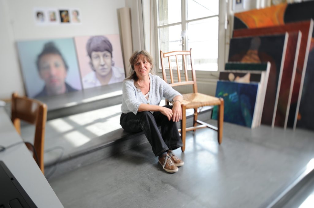 Eine Frau sitzt auf einem Absatz in ihrem Atelier. Rechts neben ihr ein Stuhl, auf welchem Sie ihren Arm abgelegt hat. Der rechte Arm ist über ihre Knie gelegt. Links im Hintergrund stehen zwei grosse Bilder an die Wand angelehnt am Boden. Beide mit einem Gesichtsporträt. Darüber sind 4 kleine Bilder angebracht. Rechts von ihr im Hintergrund ein Fenster und daneben der grösse nach sortiert, 7 Bilder exakt aufgestellt, sodass immer das vordere das hintere verdeckt.